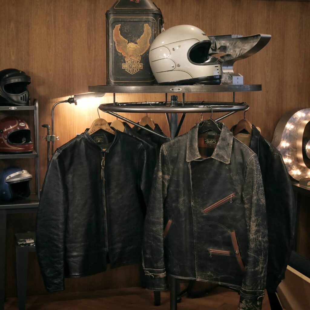 ガレージをイメージしたGANZショールームのディスプレイ。メッシュ棚オプションを装着したサークルハンガーラックにレザージャケットが掛けられ、周囲にヴィンテージヘルメット、ハーレーダビッドソンのオイル缶、背景にヘルメットラックが配置されている空間。