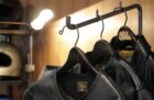 GANZの傾斜ハンガーラック「SLANT ARM RACK T-2」のアーム部分の拡大写真。黒皮鉄の傾斜アームに掛けられた3着のレザージャケットと、背景のウォルナットの木壁、温かみのある間接照明が調和したガレージ風インテリアの様子。