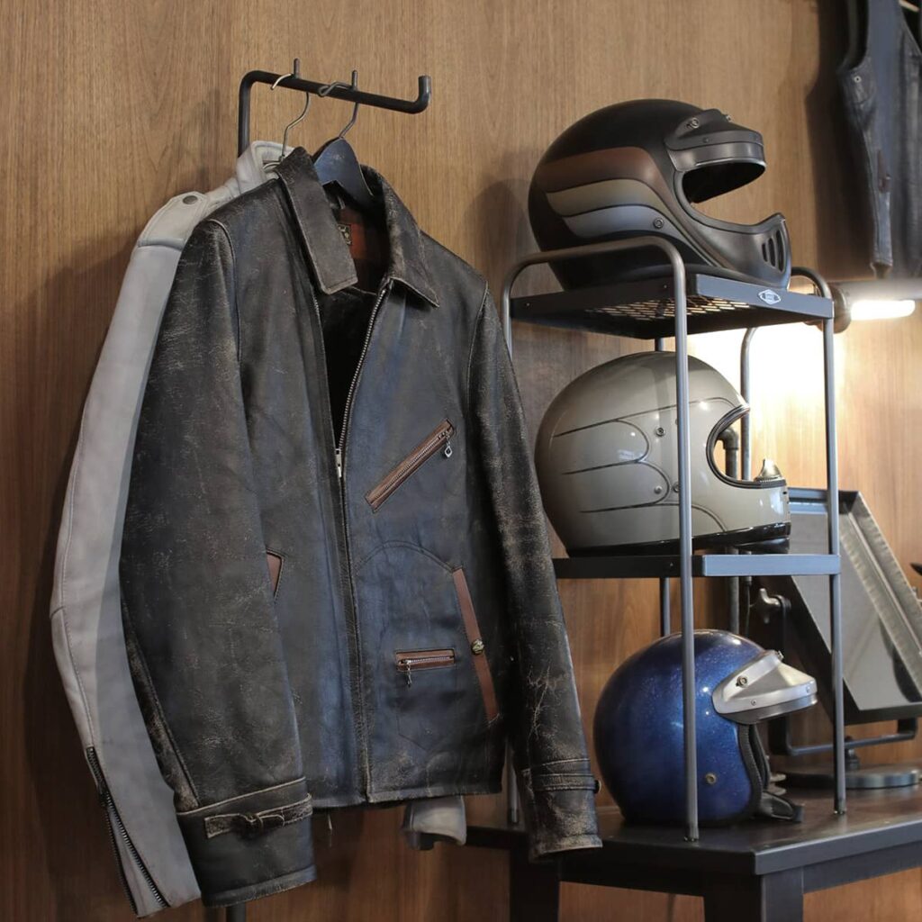 鉄製ハンガーラック「SLANT ARM RACK T-2」に掛けられた黒いライダースジャケットと、その横に並ぶGANZ製の壁掛けヘルメットラック(ビンテージヘルメットディスプレイ)。インダストリアルなガレージ空間での使用イメージ。